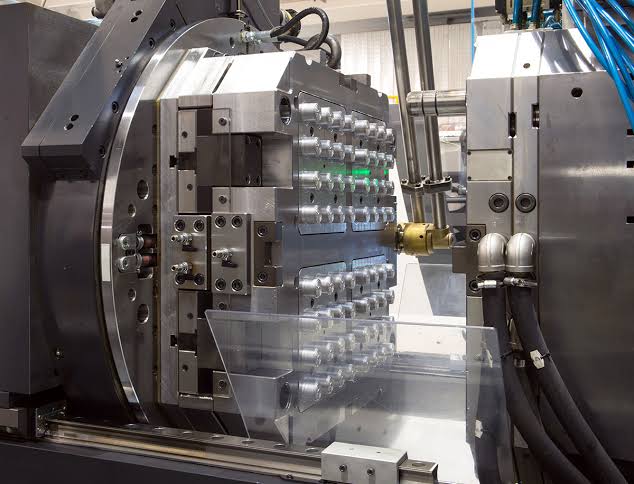 Trabajador con traje azul junto a maquinaria industrial en fábrica, con cajas apiladas y tuberías Synergy Mechatronics.